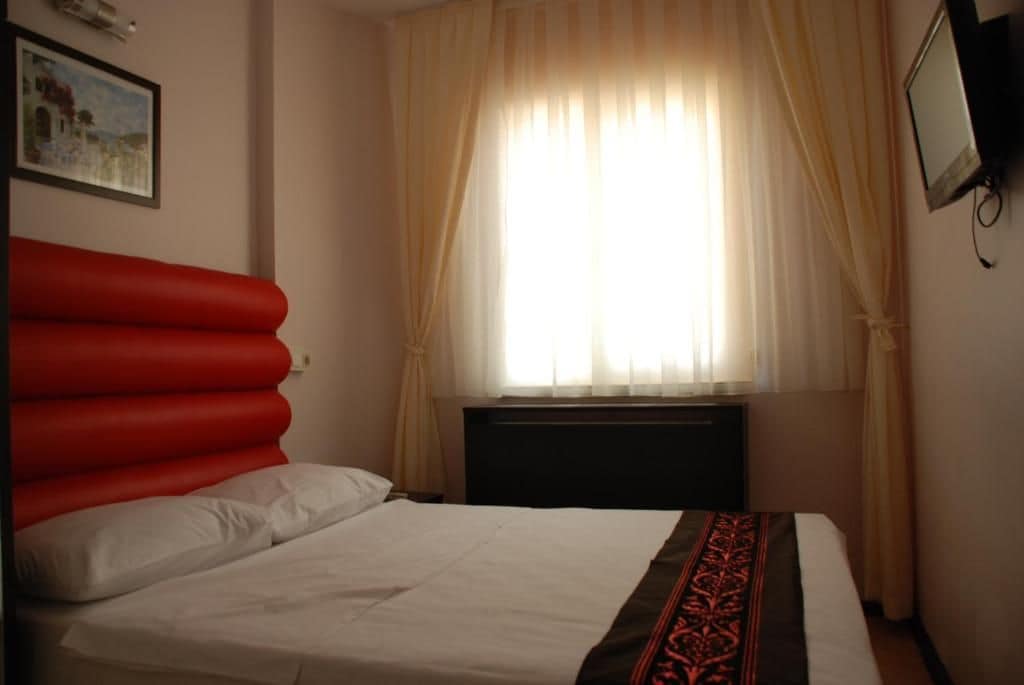 A hotel room with a bed, a window with curtains, a wall-mounted TV, and a painting.