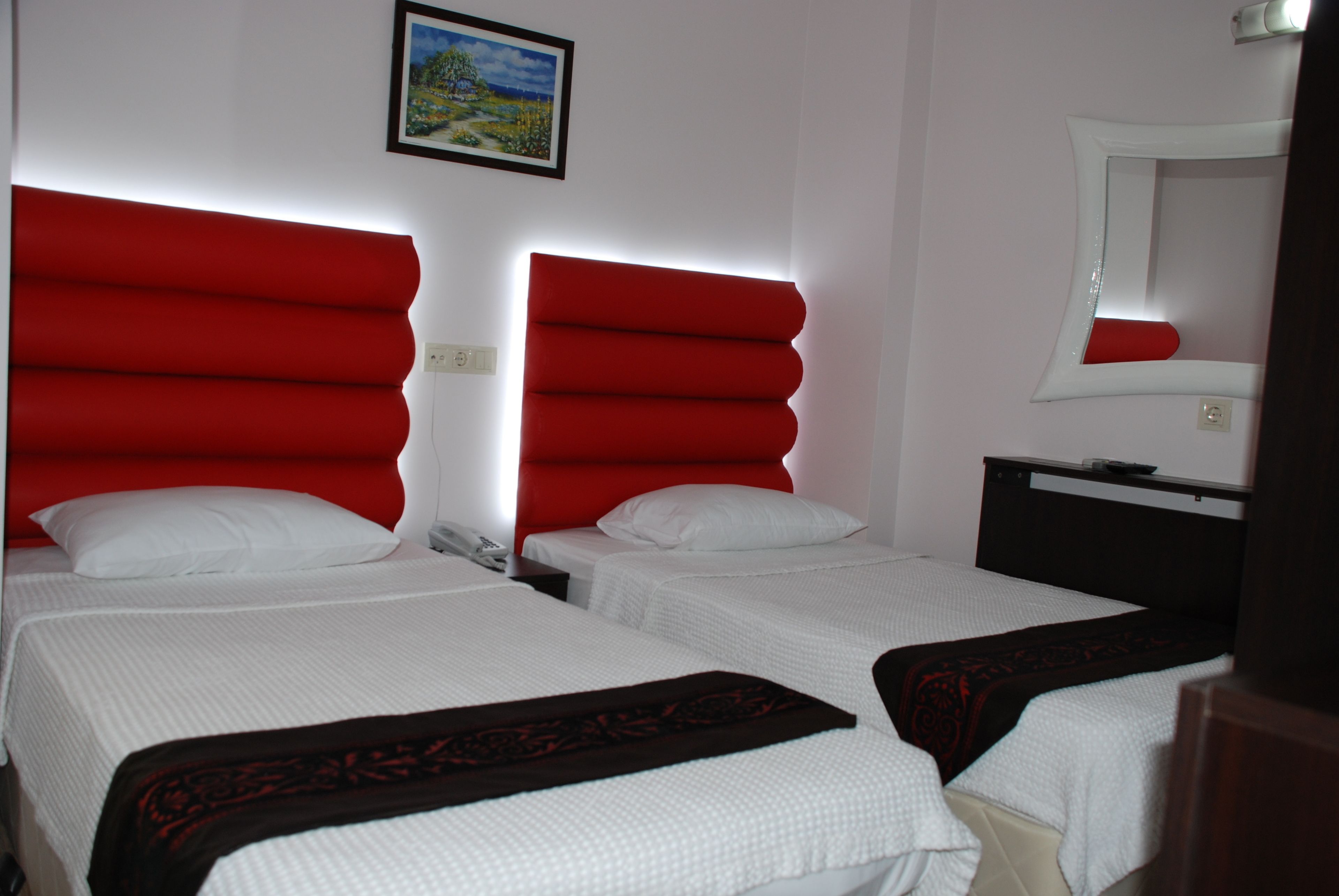 A hotel room with two beds, each with a red headboard, white bedding, and a small bedside table. A painting is visible on the wall above the beds, and a mirror is on the wall to the right.