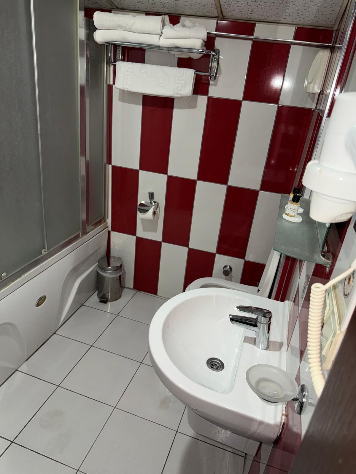 Un baño con pared de cuadros rojos y blancos, un lavamanos blanco con espejo y un perchero con toallas dobladas.