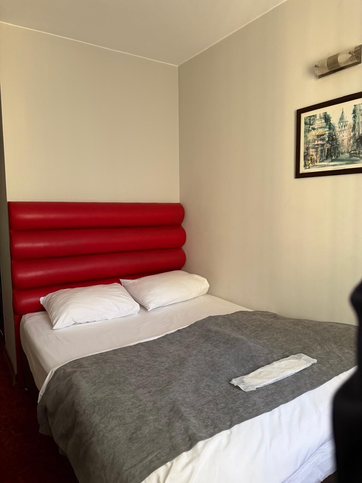 Una cama individual con cabecera roja, almohadas blancas y manta gris.