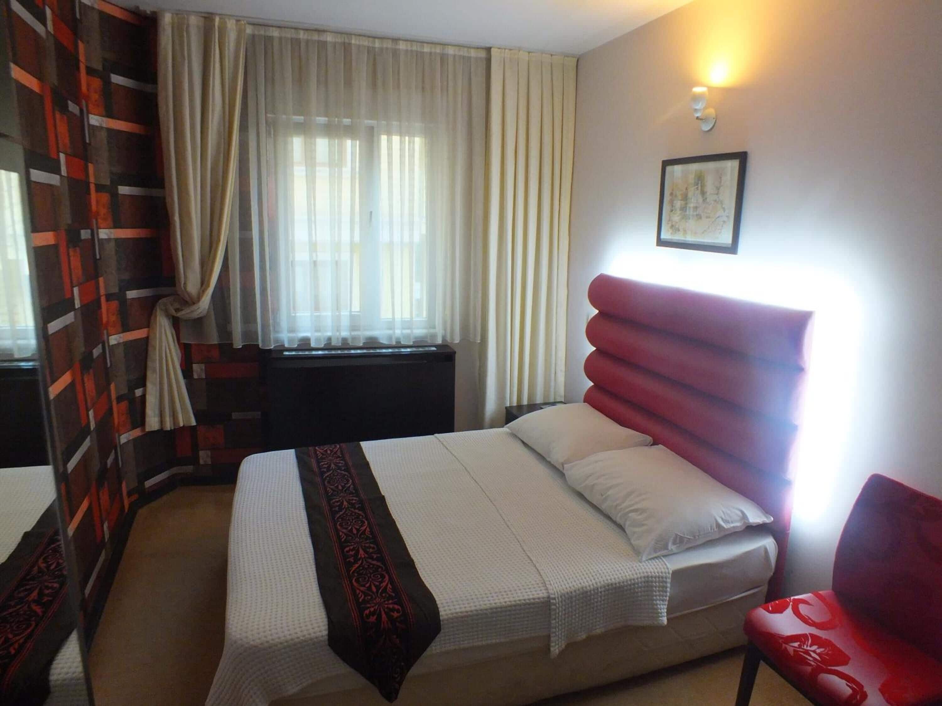 A hotel room with a bed, a red chair, a window with curtains, and a wall-mounted light.