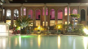 Un área de piscina con un edificio de ladrillo al fondo, que cuenta con ventanas arqueadas y una zona de estar cubierta con sombrillas.
