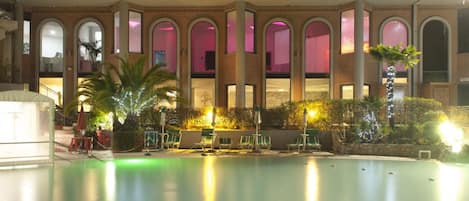 Un área de piscina con un edificio de ladrillo al fondo, que cuenta con ventanas arqueadas y una zona de estar cubierta con sombrillas.