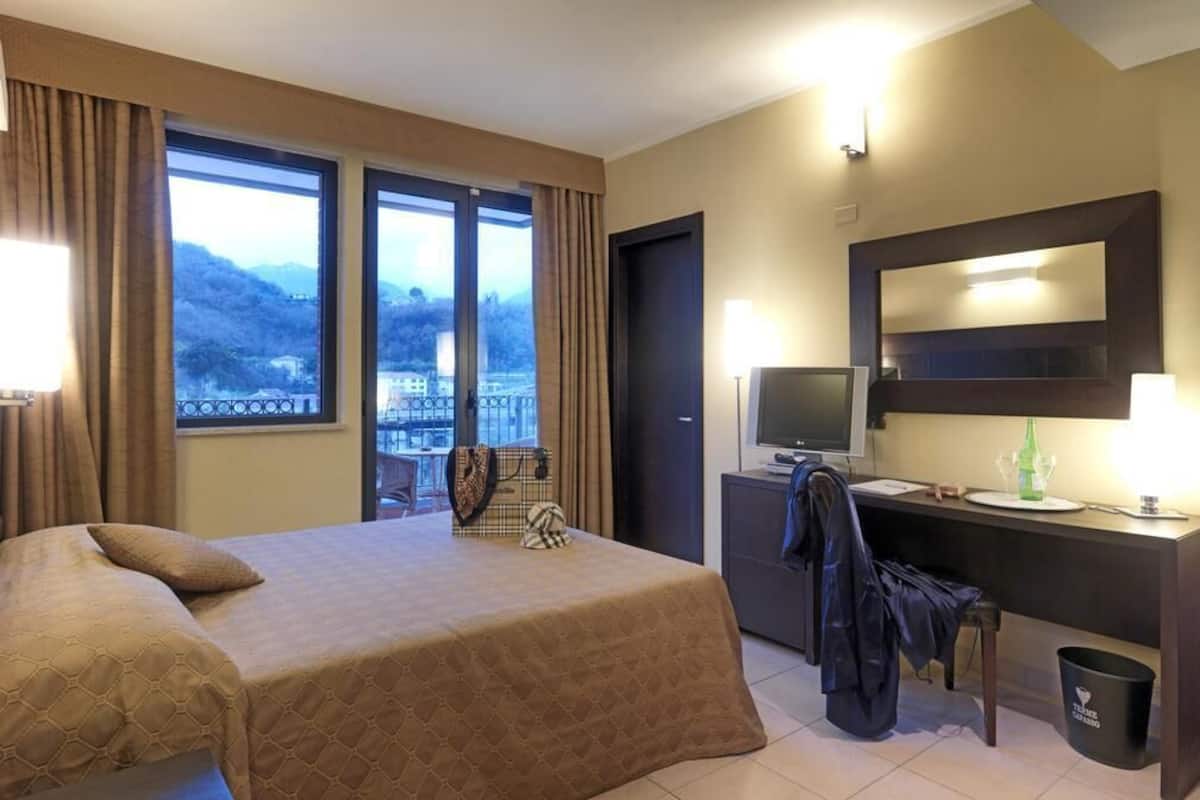 A hotel room with a bed, a desk with a TV, a chair, and a view of the mountains.