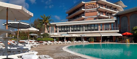 A hotel with a pool area, sun loungers, umbrellas, and a clear blue sky.