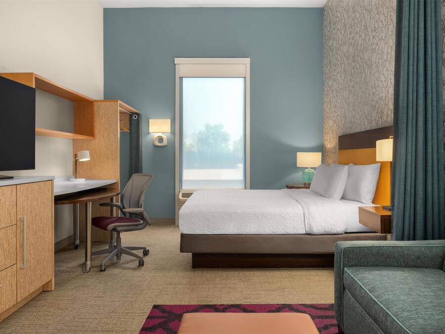 Una habitación de hotel con cama, escritorio, silla, televisor y ventana con cortinas.