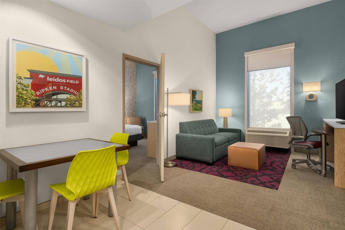 A hotel room with a dining table, chairs, a sofa, a small ottoman, a desk with a chair, a window with blinds, a lamp, and a framed picture on the wall.