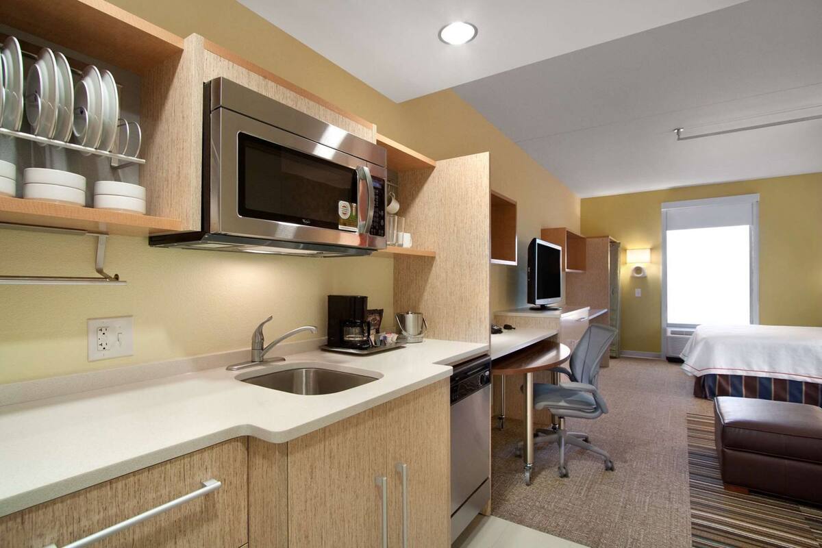 A compact hotel room with a kitchenette, a bed, a desk with a chair, and a large window.
