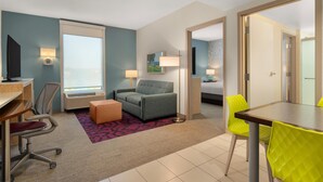 A hotel room with a desk, office chair, sofa, and a dining table with chairs. There is a window with blinds, a painting on the wall, and a door leading to another room. - Home2 Suites by Hilton Baltimore / Aberdeen, MD (Aberdeen)