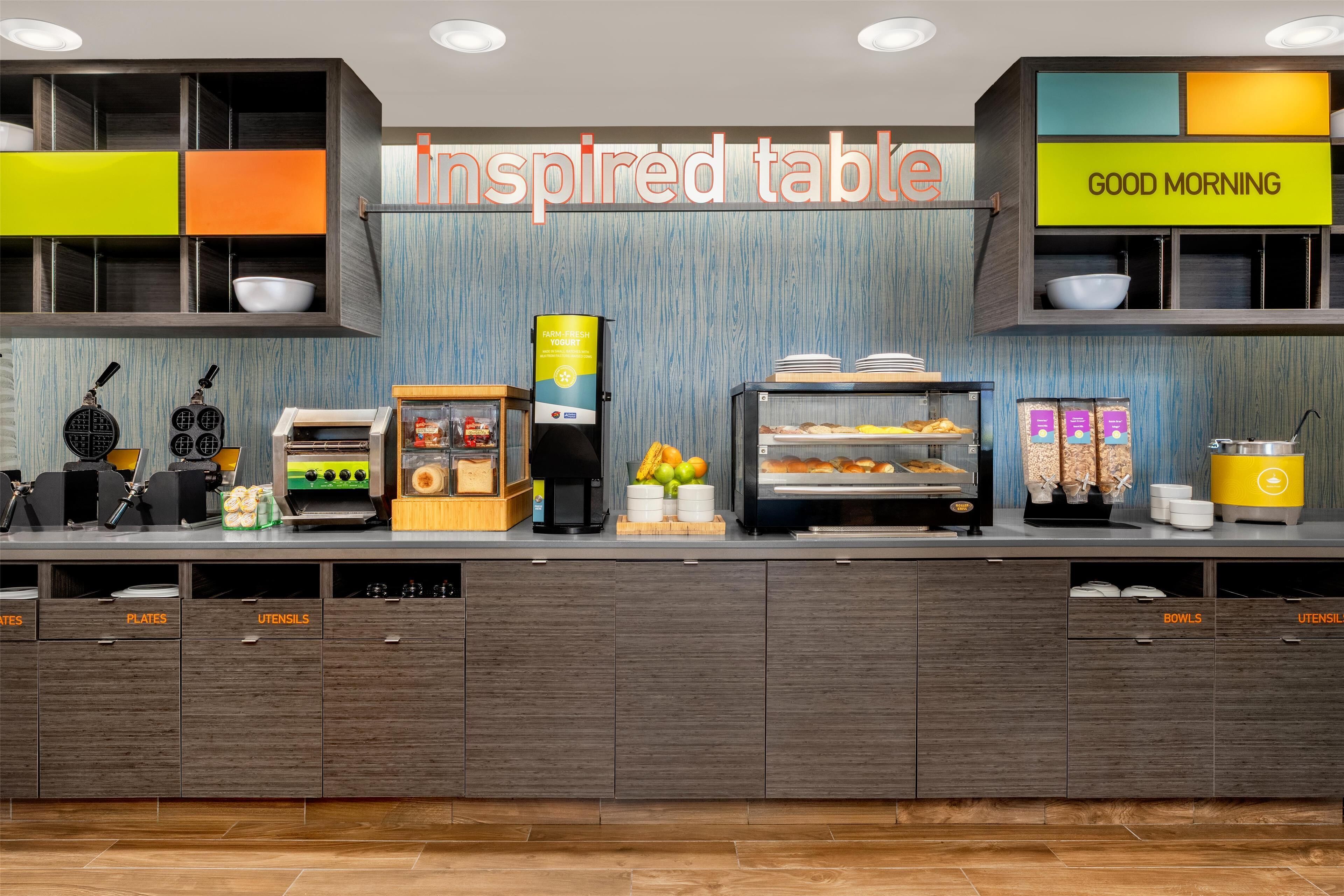 A breakfast bar with various food items and appliances, including waffle makers, a coffee machine, and a yogurt dispenser.