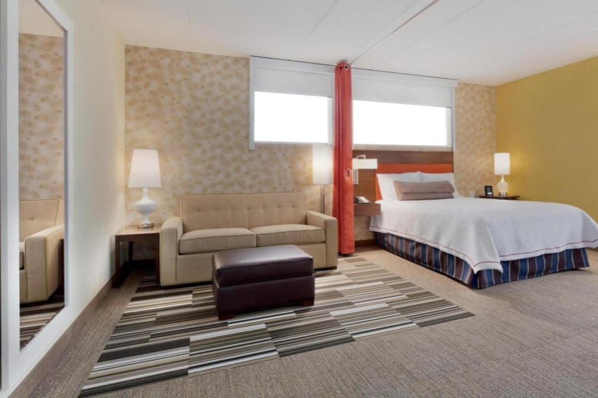 A hotel room with a bed, a sofa, a nightstand, a lamp, a window, and a mirror.