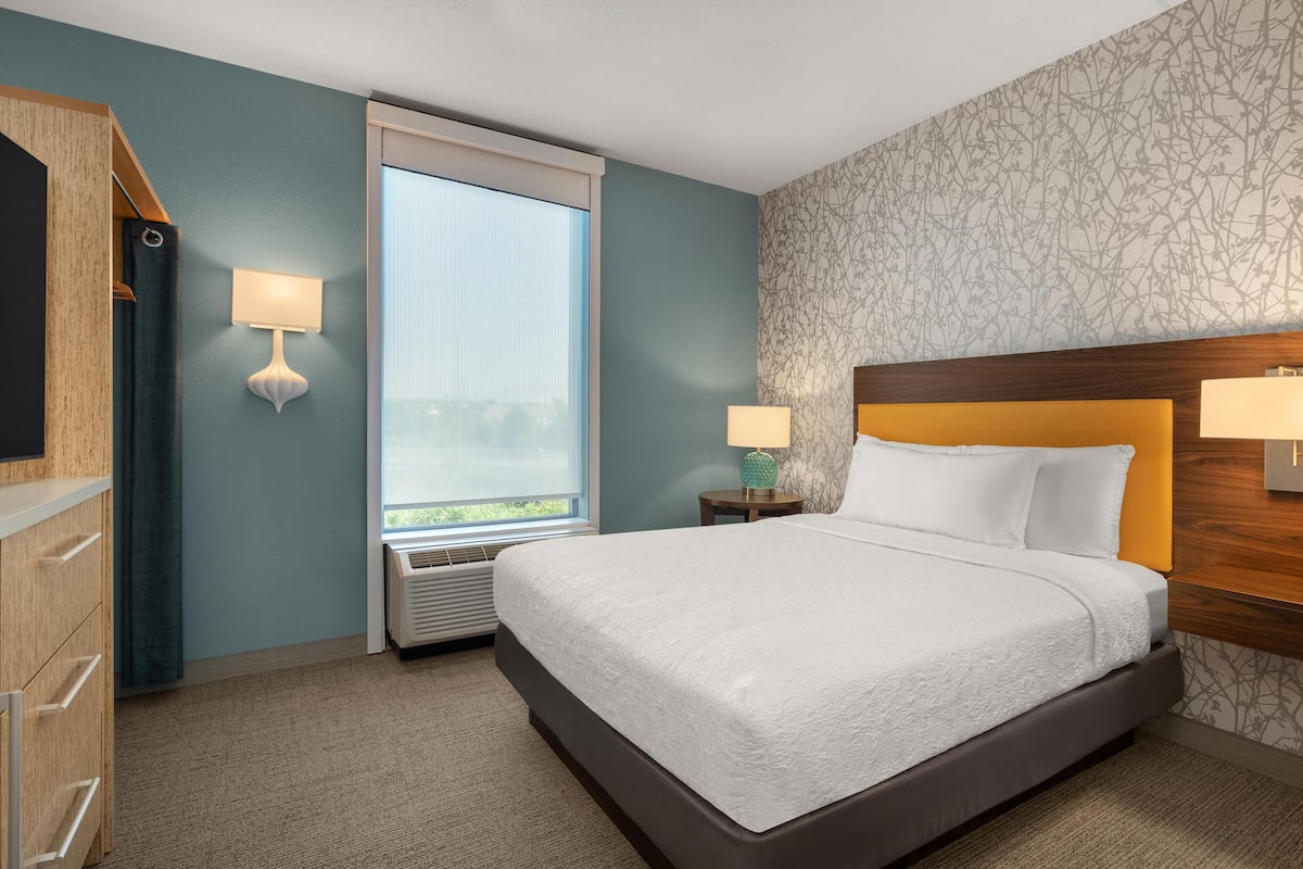A hotel room with a large bed, two bedside lamps, a window with a view, a wooden headboard, and a wall-mounted TV.