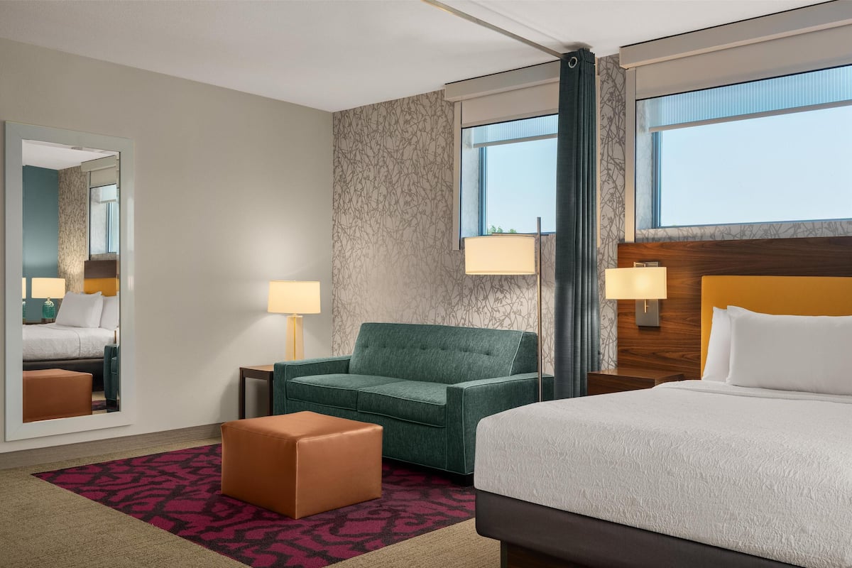 A hotel room with a bed, a green armchair, a brown ottoman, a large mirror, and a window with a view.