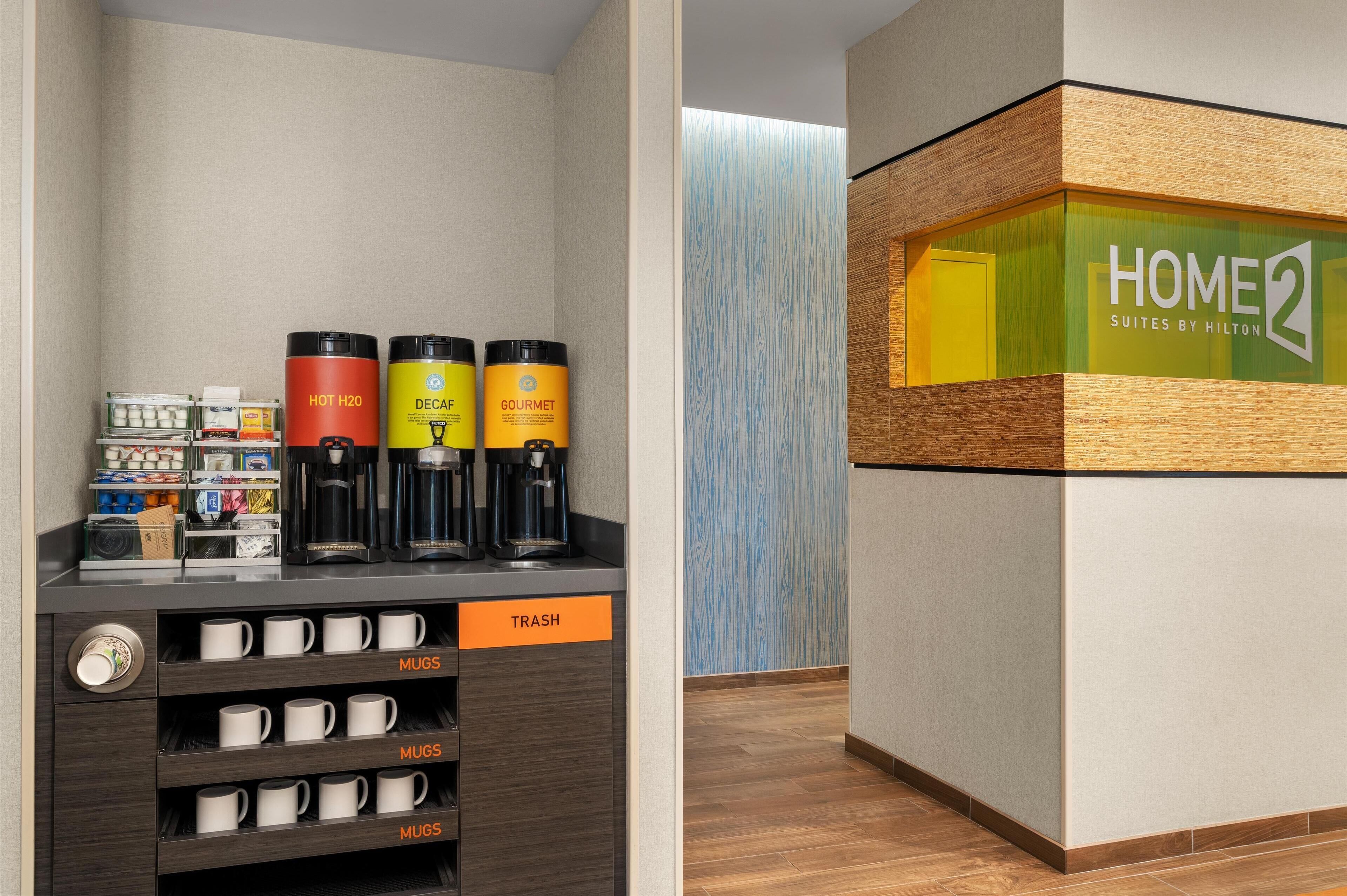 A hotel reception area with a coffee station, a trash bin, and a sign for 'HOME2 Suites by Hilton'.