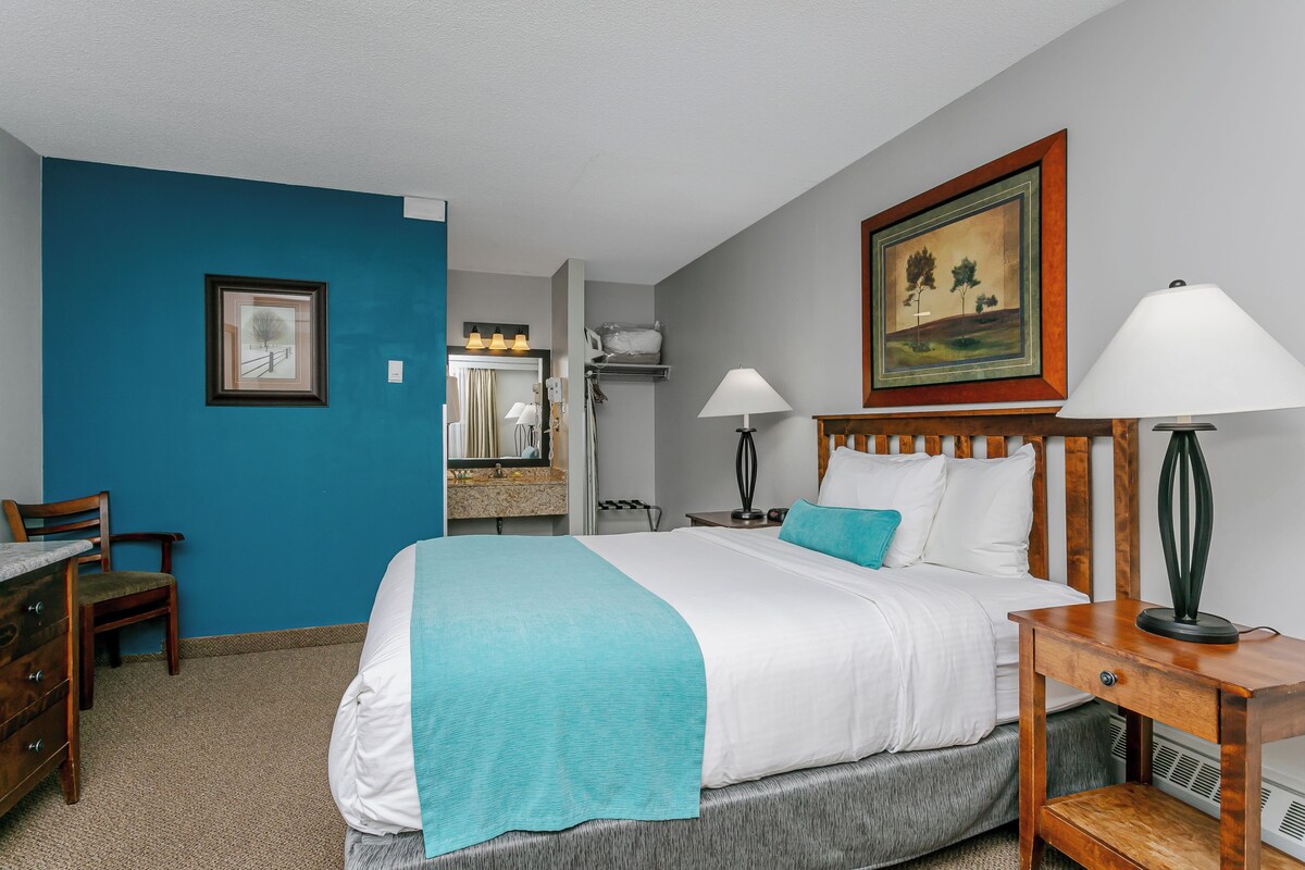 A hotel room with a bed, a desk, a chair, a bathroom, and a painting on the wall.