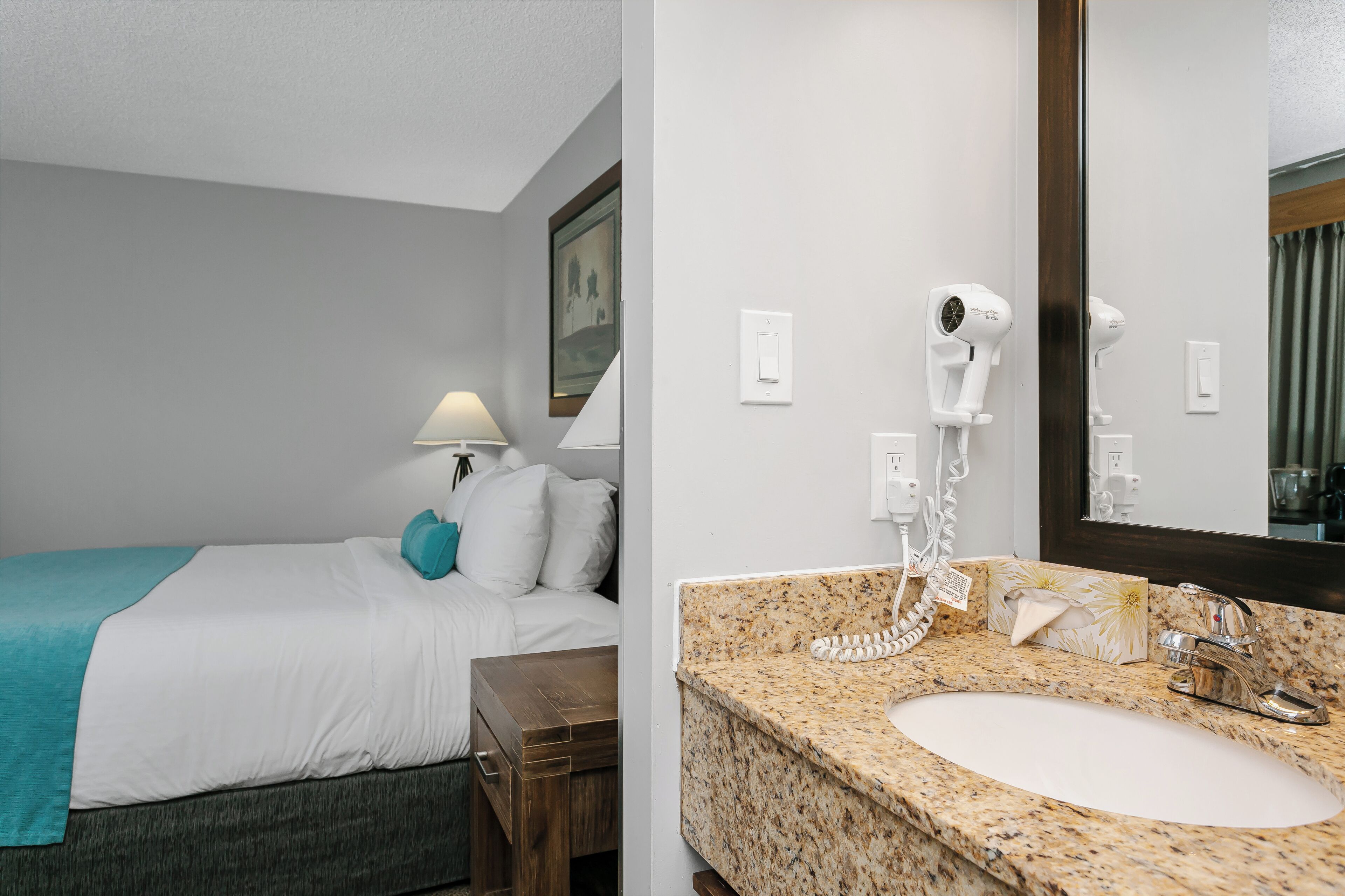 A hotel room with a bed, a nightstand, a lamp, a hairdryer, and a bathroom with a sink and mirror.