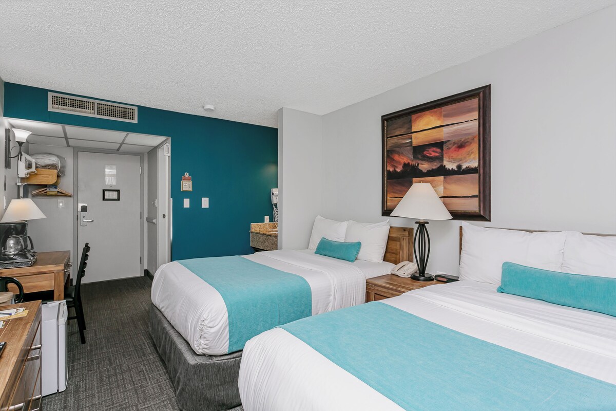 A hotel room with two beds, a desk, a chair, a coffee maker, and a painting on the wall.