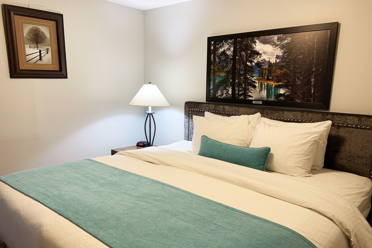 A neatly made bed with a green throw, a bedside lamp, a clock, and a framed picture on the wall.