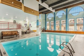 An indoor swimming pool with a wooden bench, a red railing, and large windows with reflections of the outside.