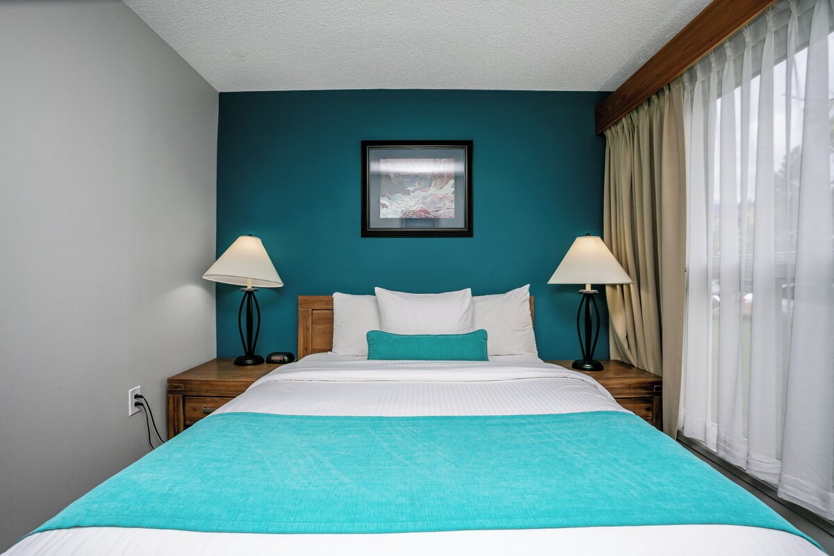 A hotel room with a bed, two bedside tables, a nightstand, a framed picture on the wall, and a window with curtains.
