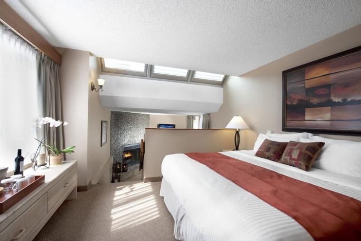 A hotel room with a large bed, a desk, a chair, a small table, a lamp, and a painting on the wall.