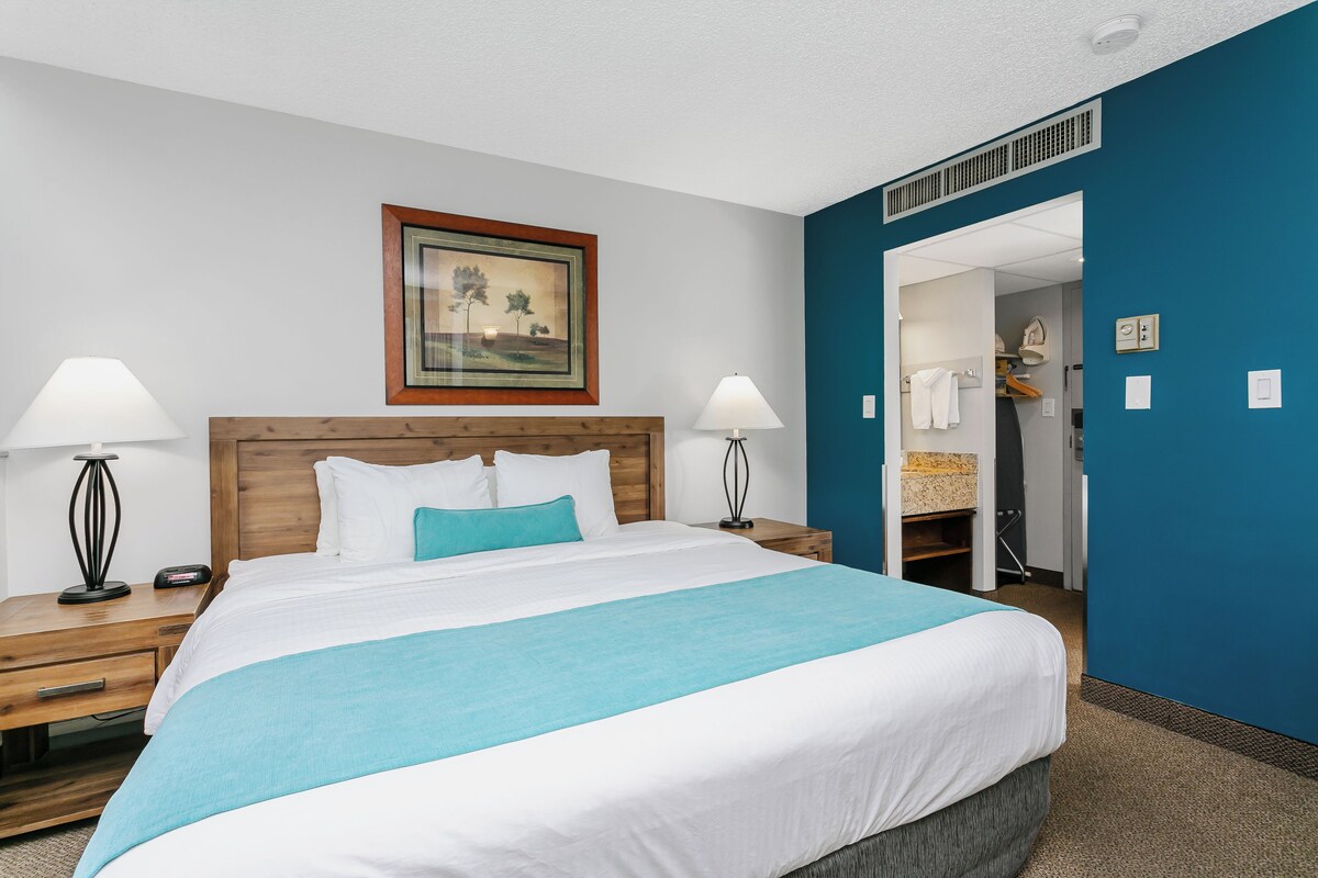 A hotel room with a large bed, two bedside tables with lamps, a framed picture on the wall, and a bathroom visible through an open door.