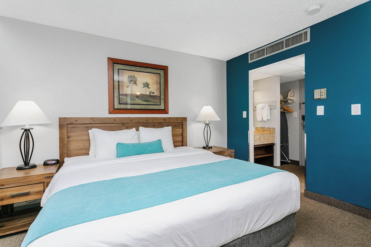 A hotel room with a large bed, two bedside tables with lamps, a framed picture on the wall, and a bathroom visible through an open door.