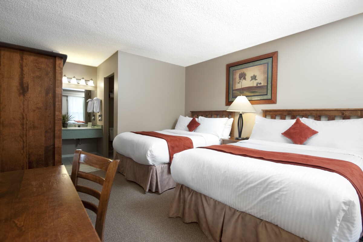 A hotel room with two beds, a wooden desk, a mirror, and a painting on the wall.