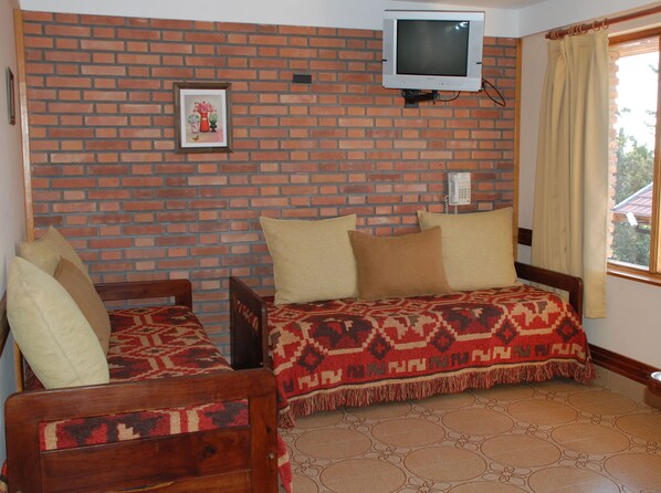 A room with a brick wall, a television mounted above a telephone, a bed with patterned bedding, and a window with curtains.