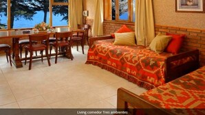 A cozy room with a bed, a dining table, and chairs. There are large windows with curtains, and a painting on the wall.