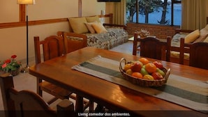 A dining area with a wooden table, chairs, and a fruit basket. A sofa and a television are visible in the background.