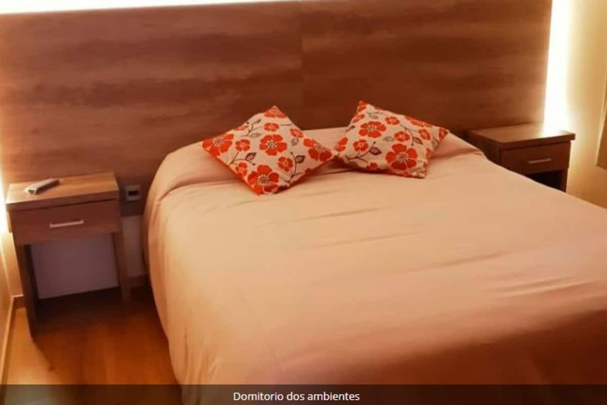 Ein Schlafzimmer mit einem hölzernen Kopfteil, einem Bett mit einer beige farbenen Tagesdecke, zwei Nachttischen, wobei einer einen Fernseher mit Fernbedienung hat, und einer Schublade im linken Nachttisch.