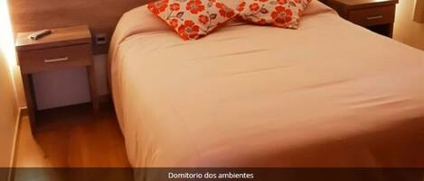 Une chambre à coucher avec une tête de lit en bois, un lit recouvert d’une couvre-lit beige, deux tables de chevet, dont l’une est surmontée d’une télécommande et l’autre, à gauche, d’un tiroir.