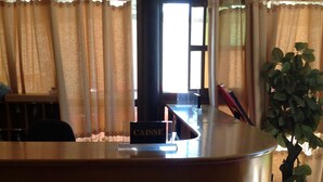 A reception desk with a sign reading "CAISSE," a potted plant, and a window with curtains. - Nikki Hotel (Niamey)