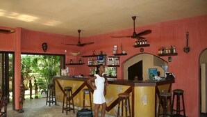 A bar area with a woman standing, bar stools, and a counter with various bottles. - Hôtel Aurore (Lomé)