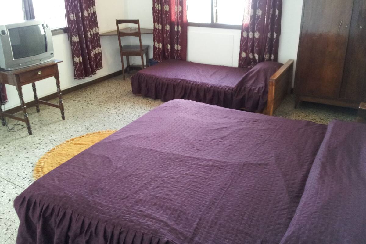 A room with two beds, a wooden chair, a small table with a television, and a window with curtains.