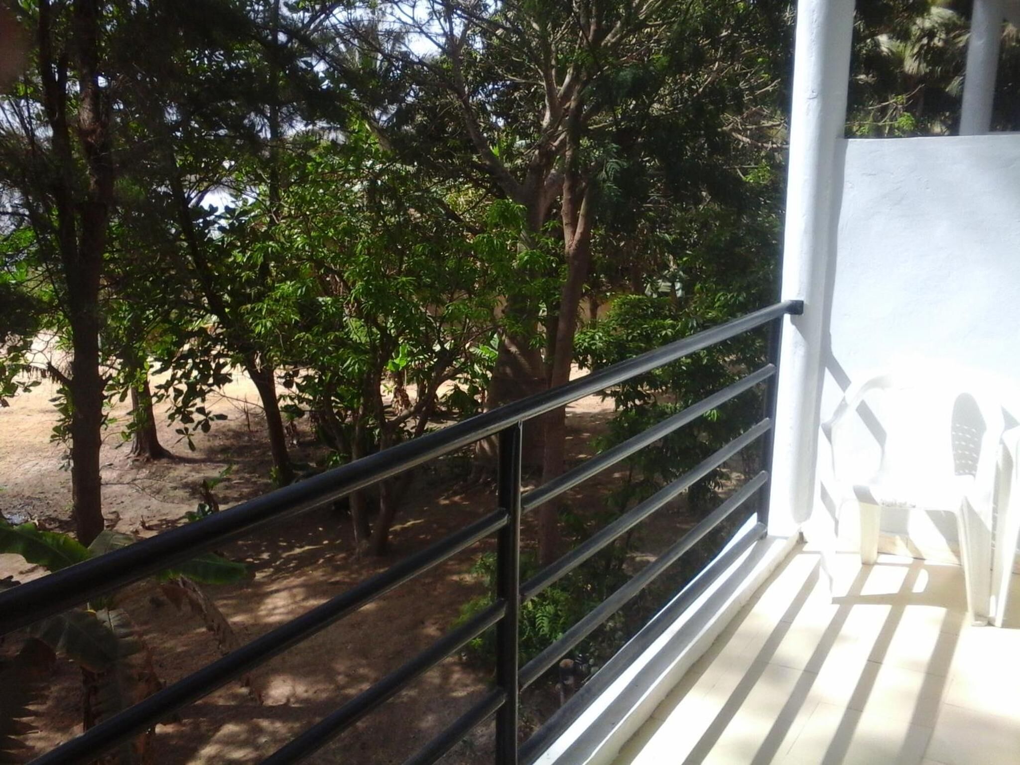 A balcony with a metal railing overlooking a garden with trees and a sandy ground.