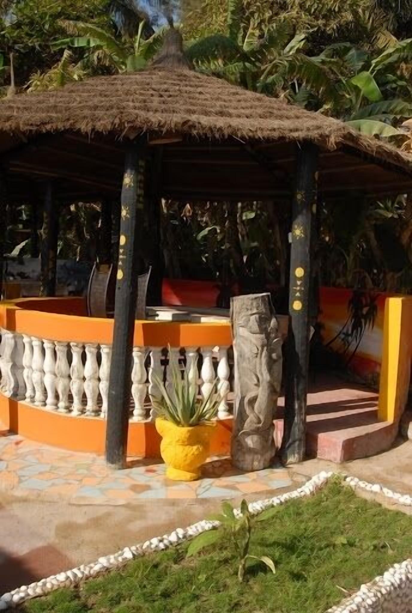 Un pabellón con techo de paja y barandilla blanca, rodeado por un patio empedrado y un pequeño jardín con una maceta amarilla y una escultura de piedra.
