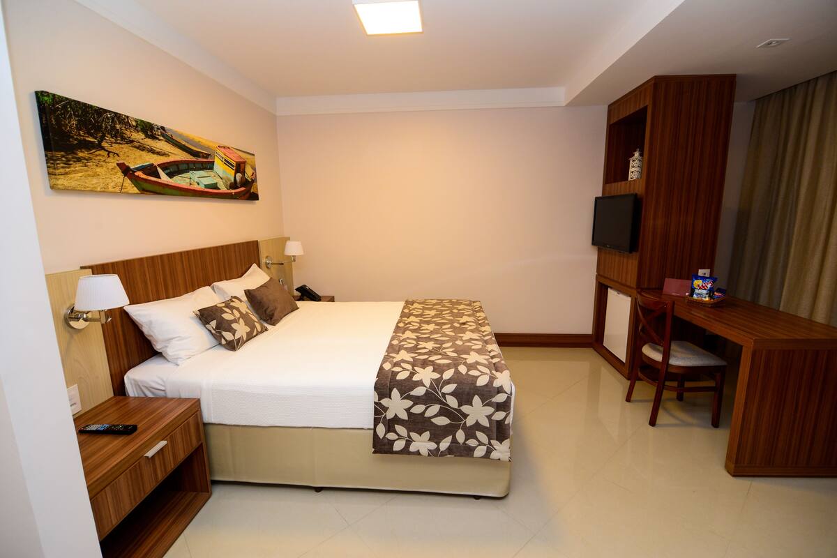 Een hotelkamer met een bed, nachtkastje, lamp, televisie, bureau, stoel en een schilderij aan de muur.