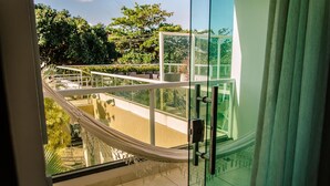 Een balkon met een hangmat, glazen hekwerk en uitzicht op groen en een gebouw.