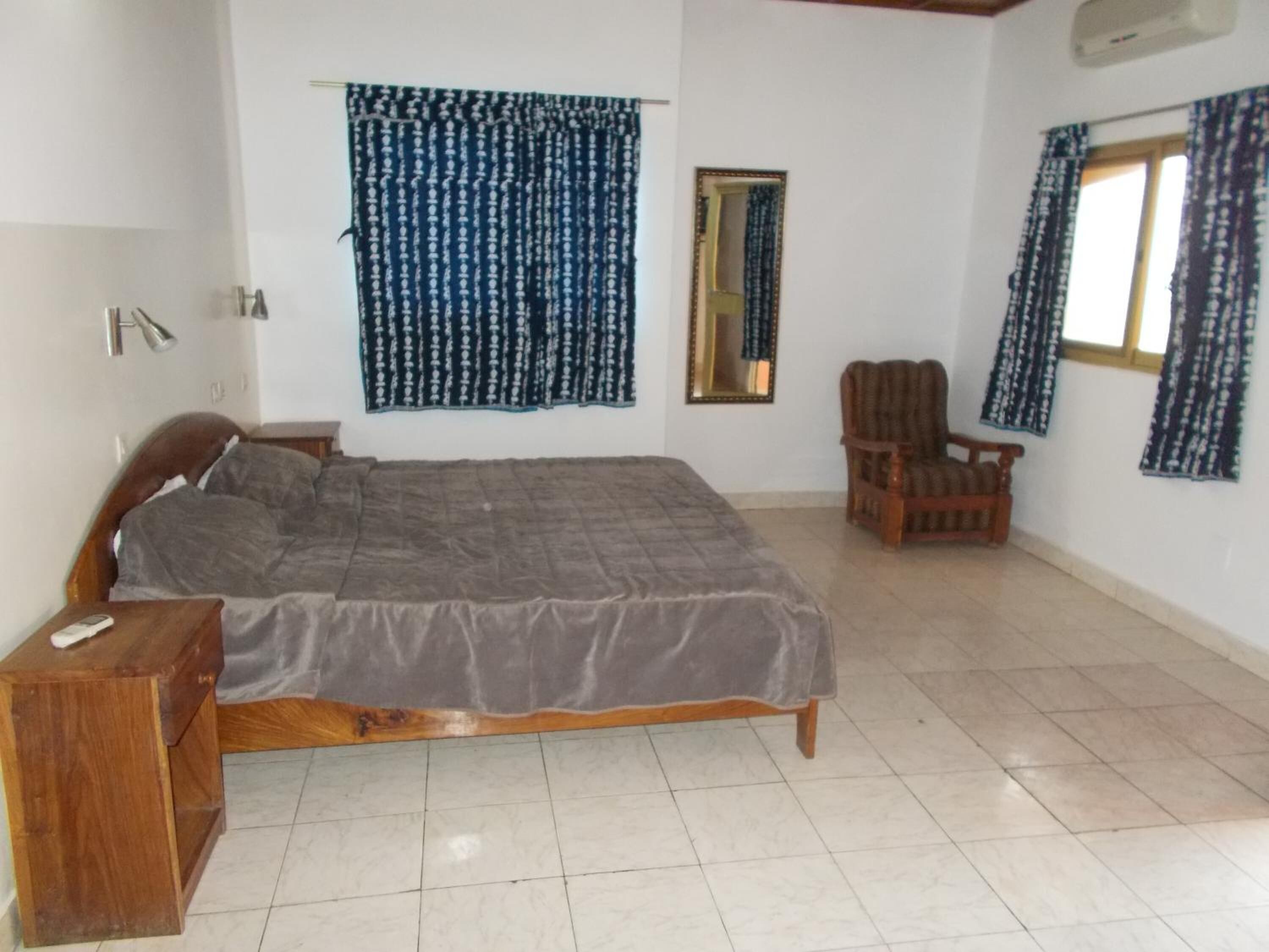 Un dormitorio con cama, mesita de noche de madera, una silla, un espejo y una ventana con cortinas.