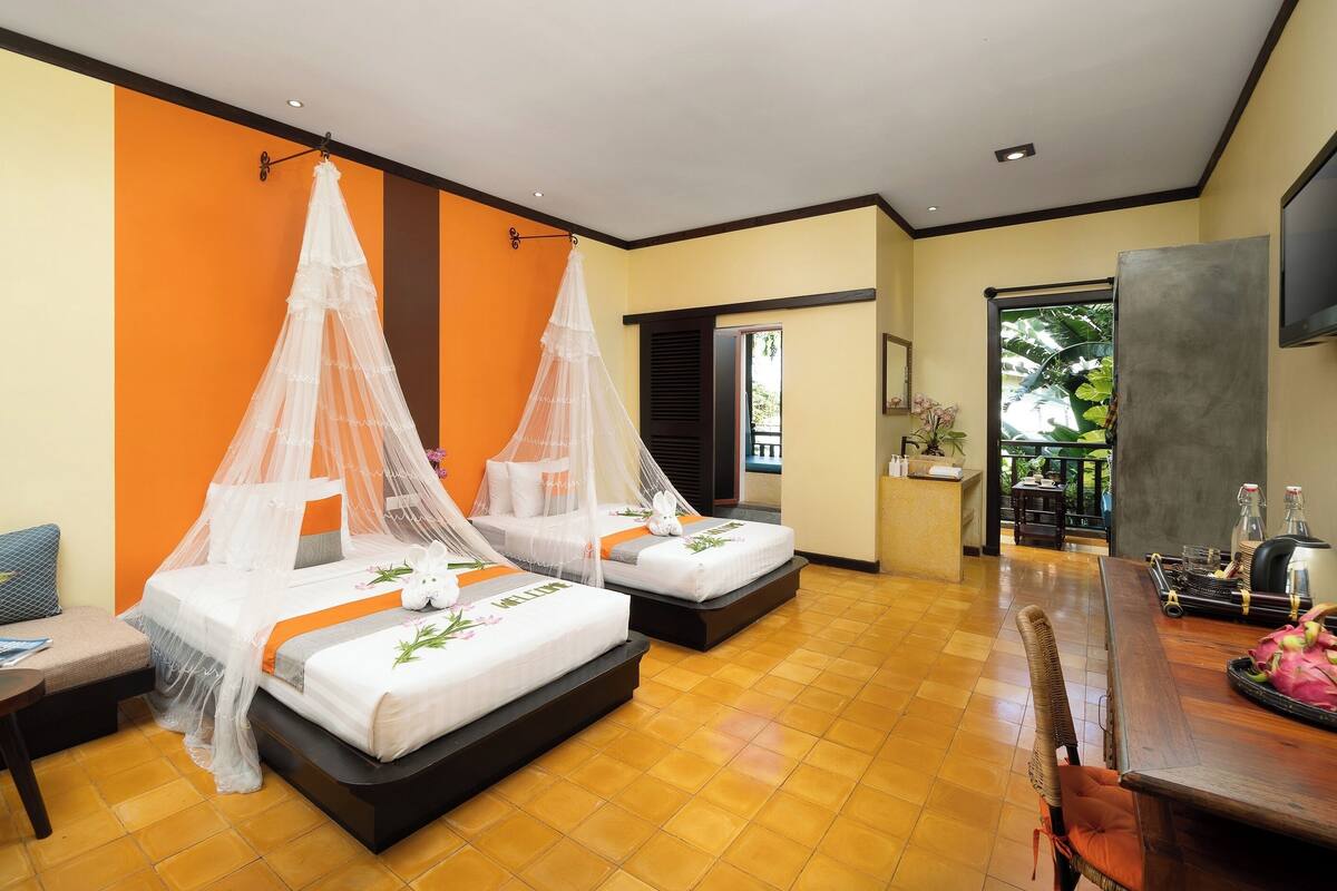 Ein Hotelzimmer mit zwei Betten, einem kleinen Tisch, einem Stuhl, einem Fernseher und einem Balkon mit Pflanzen.
