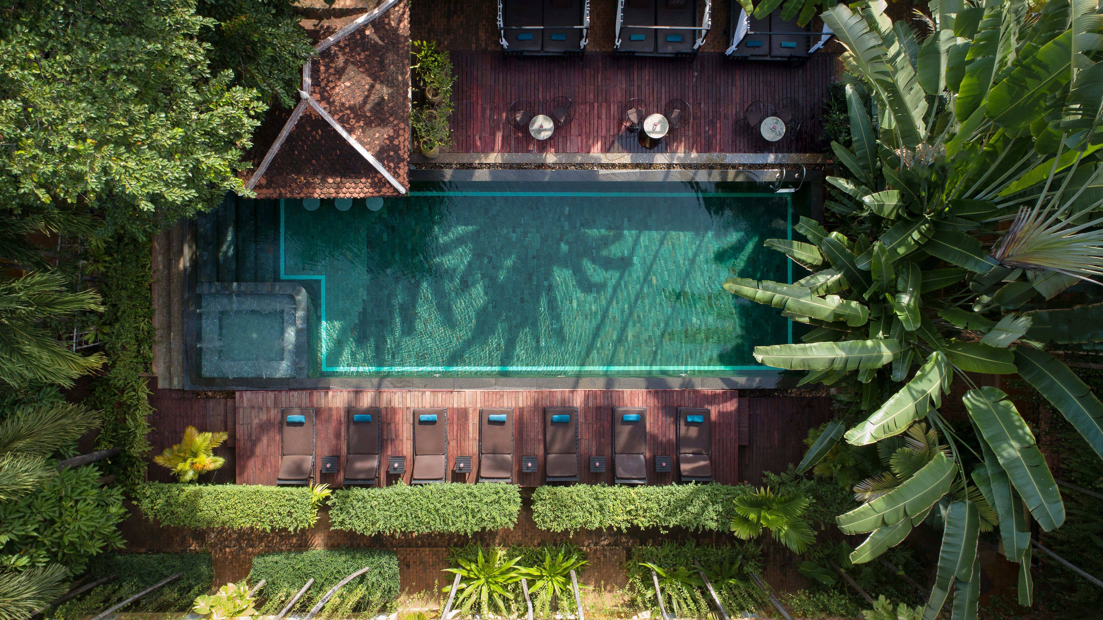 An aerial view of a pool area with a wooden deck, lounge chairs, and surrounding greenery.