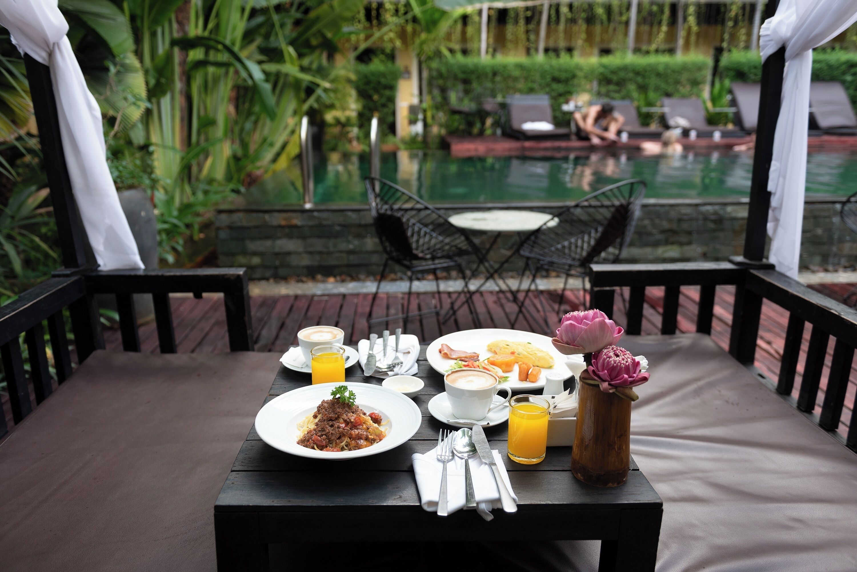 Ein Esstisch mit Frühstücksangeboten, eine Vase mit Blumen und Blick auf Pool und Liegestühle.