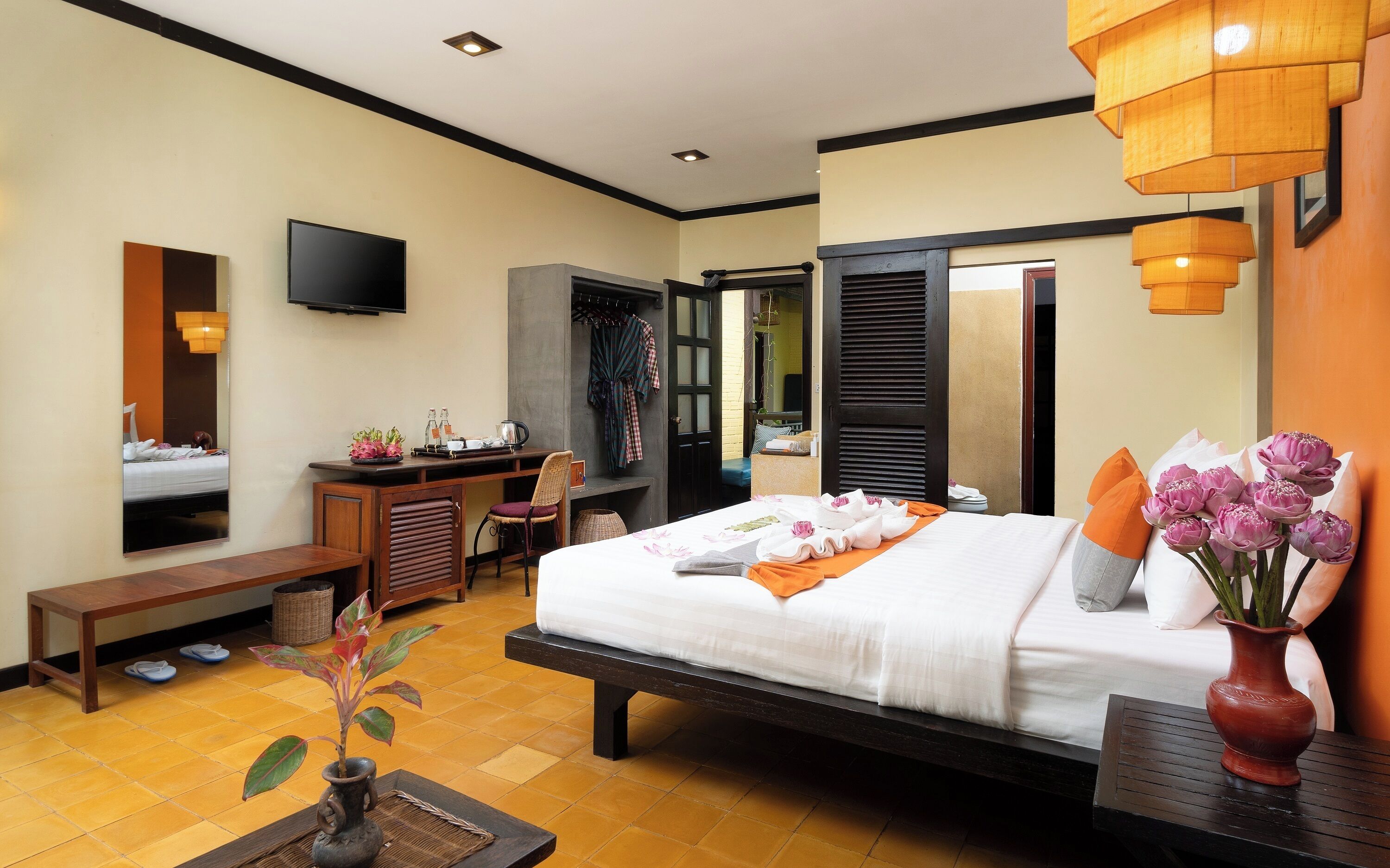A hotel room with a bed, a desk, a TV, a mirror, a bench, and a vase with flowers.