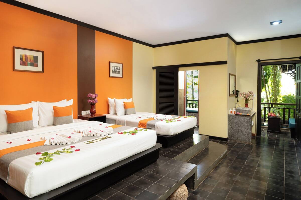 Ein Hotelzimmer mit zwei Betten, orangefarbenen Wänden, einem dunklen Boden und einem Balkon mit Pflanzen.