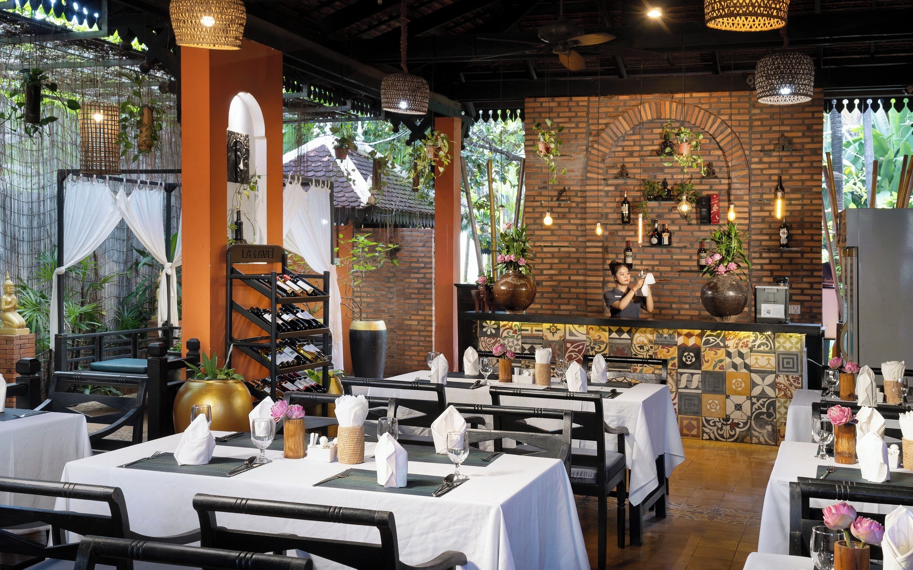 A restaurant with brick walls, hanging plants, and a wine rack. Tables are set with white tablecloths and napkins.
