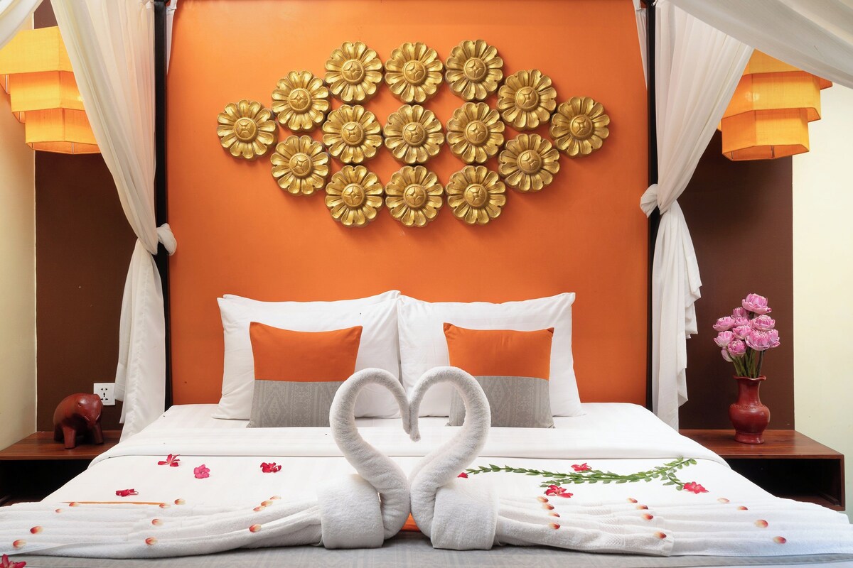 Una habitación de hotel con una cama, detalles decorativos en la pared, una toalla dispuesta en forma de corazón y un jarrón con flores.
