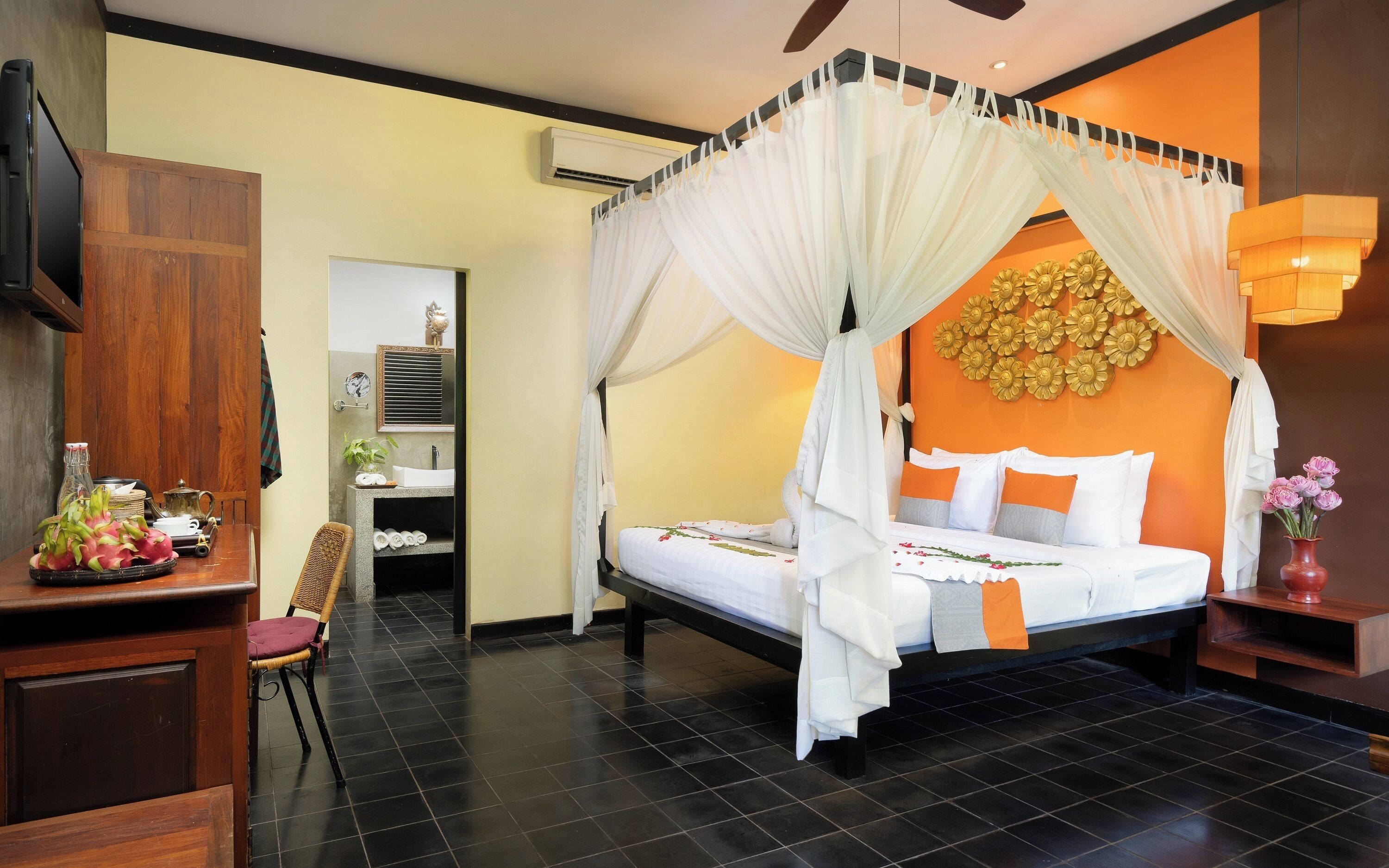 Habitación de hotel con una cama bajo un dosel, una mesita de noche con un florero y una pared decorada con flores.