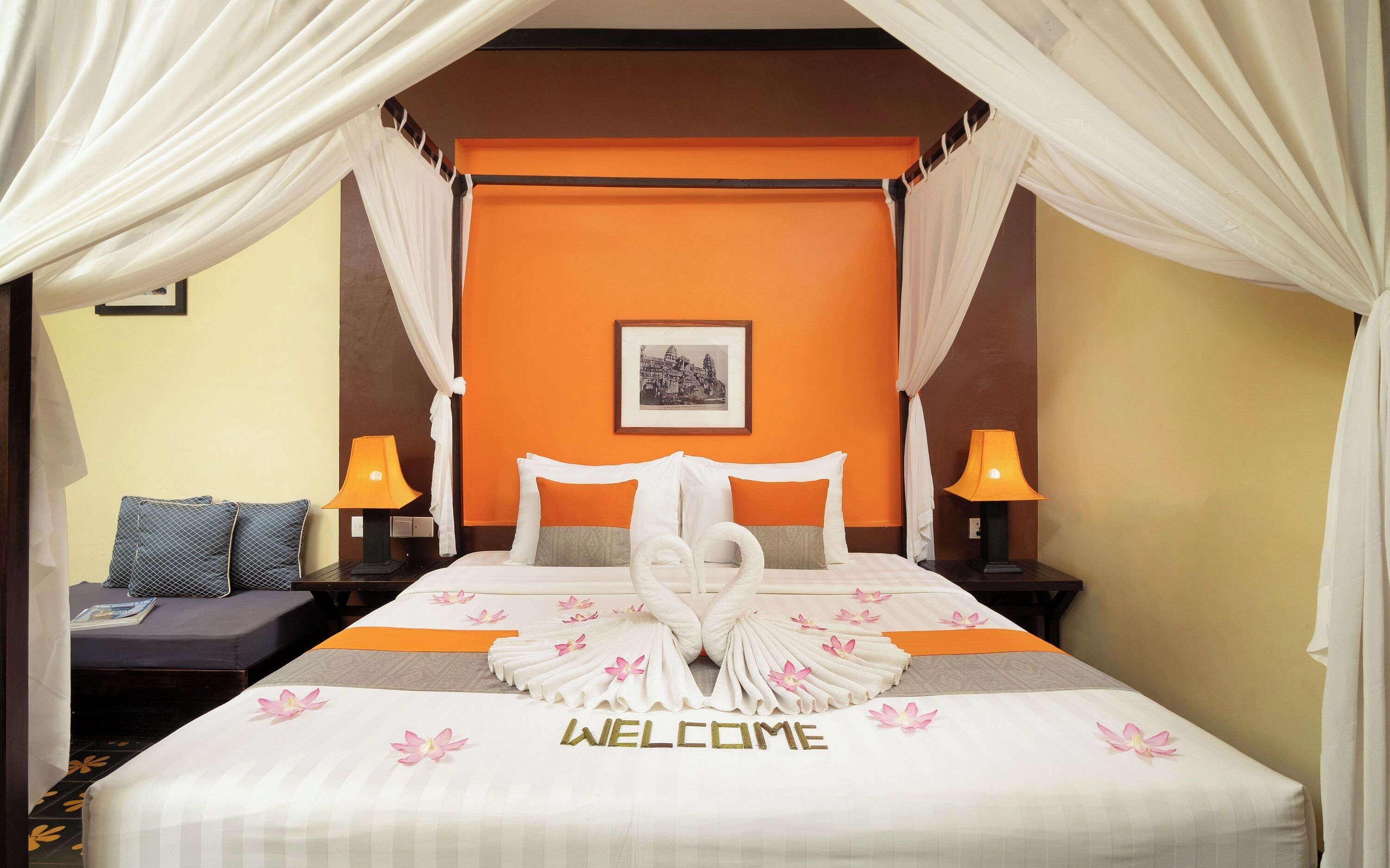 A hotel room with a bed, two nightstands, a picture on the wall, and a 'WELCOME' sign on the bed.